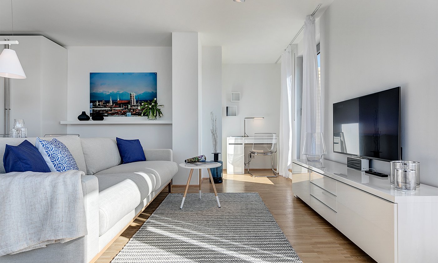 Das Bild zeigt ein Wohnzimmer mit einem weißen Sofa vor einem Fernseher. An der Wand hängt ein Bild mit der Skyline von München.